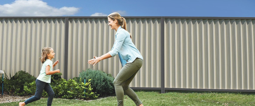 Parent and Child playing on grass in front of a grey and white Colorbond fence.